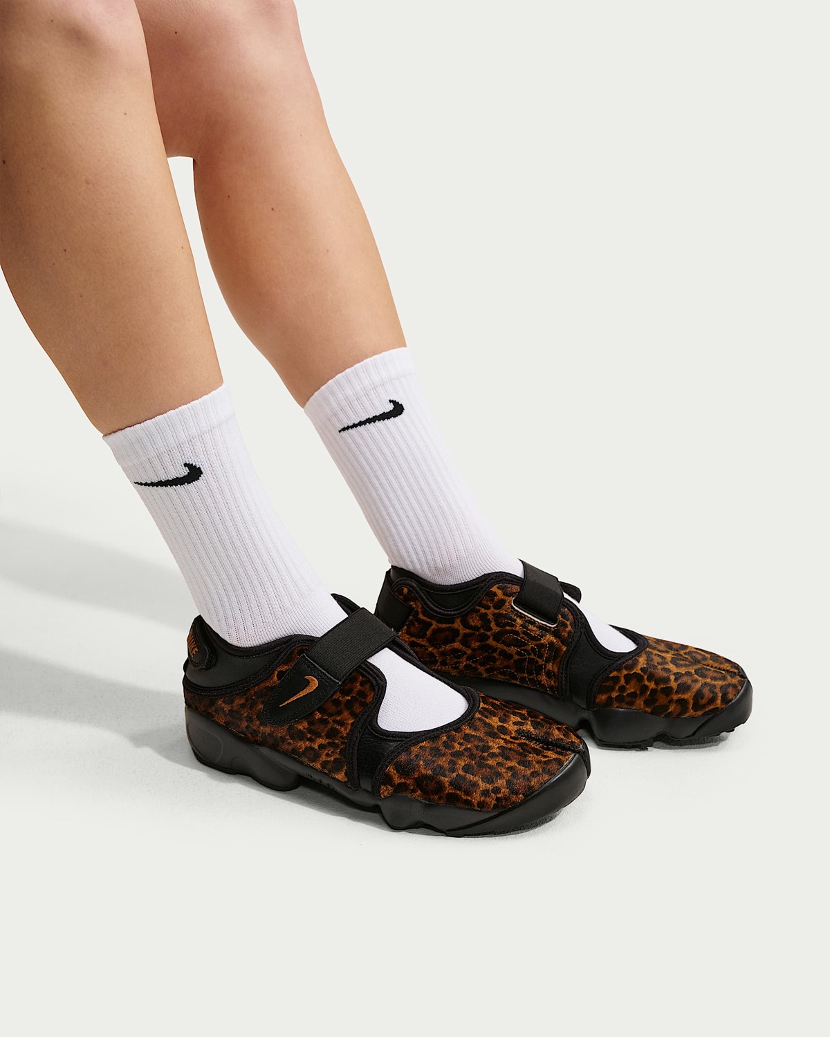 Piernas de una persona con medias blancas Nike y el par del Nike Air Rift Black/Desert Ochre puesto, con el pelo ocre y manchas negras, strap negro y suela negra, sobre un fondo claro