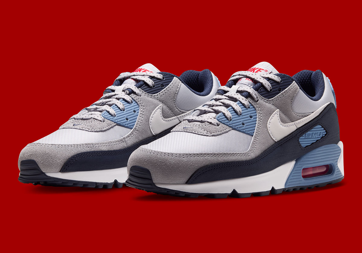 Par de Nike Air Max 90 Geyser Grey Work Blue en diagonal sobre fondo rojo, con malla gris clara, gamuza gris, Swoosh blanco, puntera azul marino, detalles Work Blue en la mediasuela y aire rojo a la vista