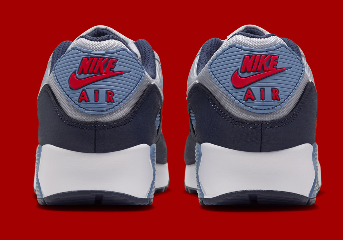 Talones del Nike Air Max 90 Geyser Grey Work Blue sobre fondo rojo, con el parche Work Blue con el logo Nike Air rojo, la base azul marino y la mediasuela blanca