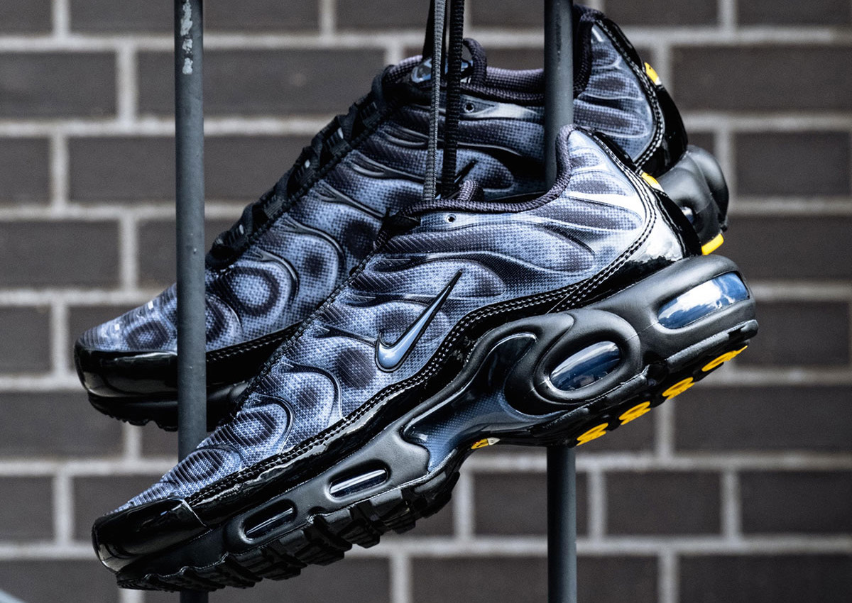 Par de Nike Air Max Plus negro y gris colgado de una baranda frente a una pared de ladrillos, con el upper en degradé de puntos, las ondas laterales, la burbuja de aire azulada y detalles amarillos en la suela