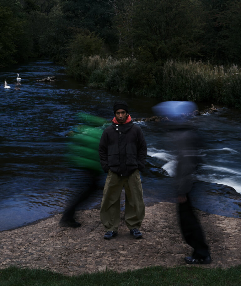 Foto de campaña de la ASICS Utility Collection: un chico con gorro negro, campera negra y pantalón cargo verde oliva parado junto a un río al atardecer, con zapatillas ASICS oscuras, mientras dos figuras pasan movidas a los costados