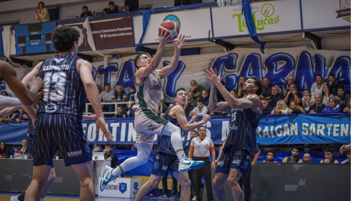 Un jugador de Gimnasia con camiseta blanca y verde salta hacia el aro entre jugadores de Argentino con camiseta azul a rayas, en El Fortín de las Morochas