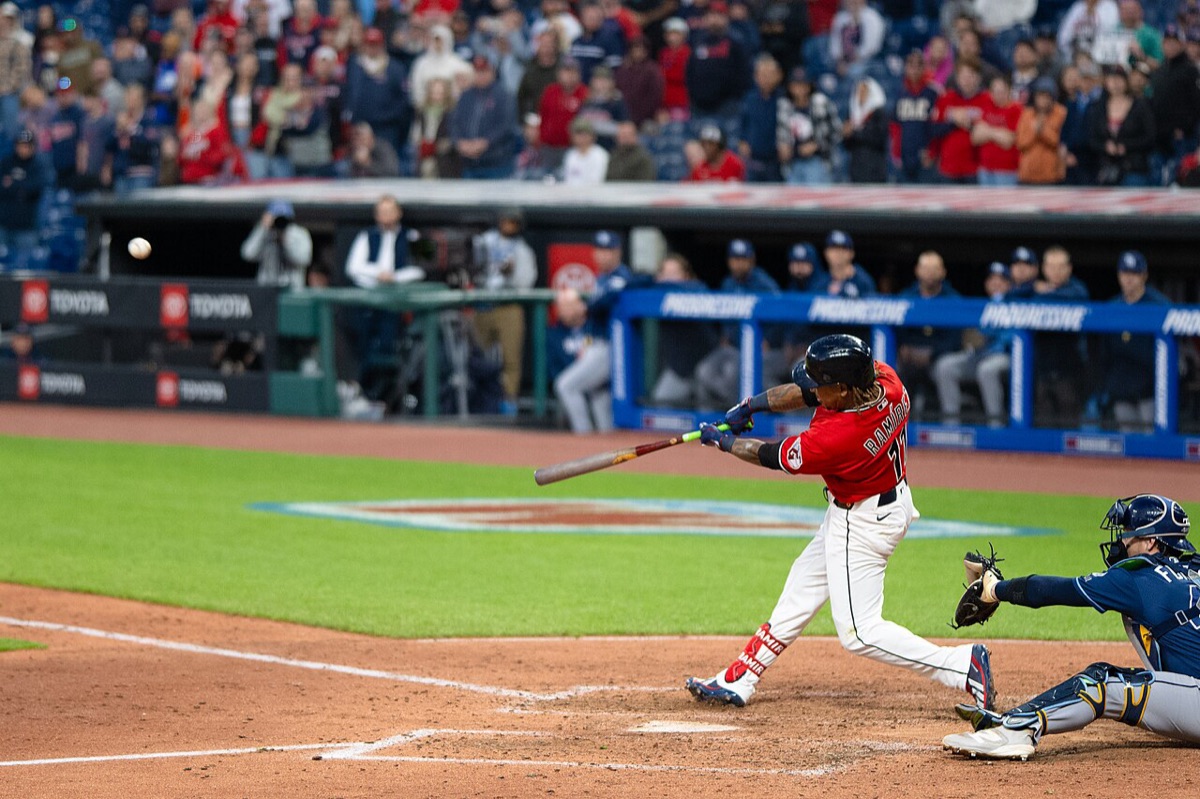 Foto de archivo: José Ramírez, de los Cleveland Guardians, con camiseta roja número 11, haciendo contacto con la pelota en el plato, con el catcher rival agachado detrás y el dugout lleno al fondo