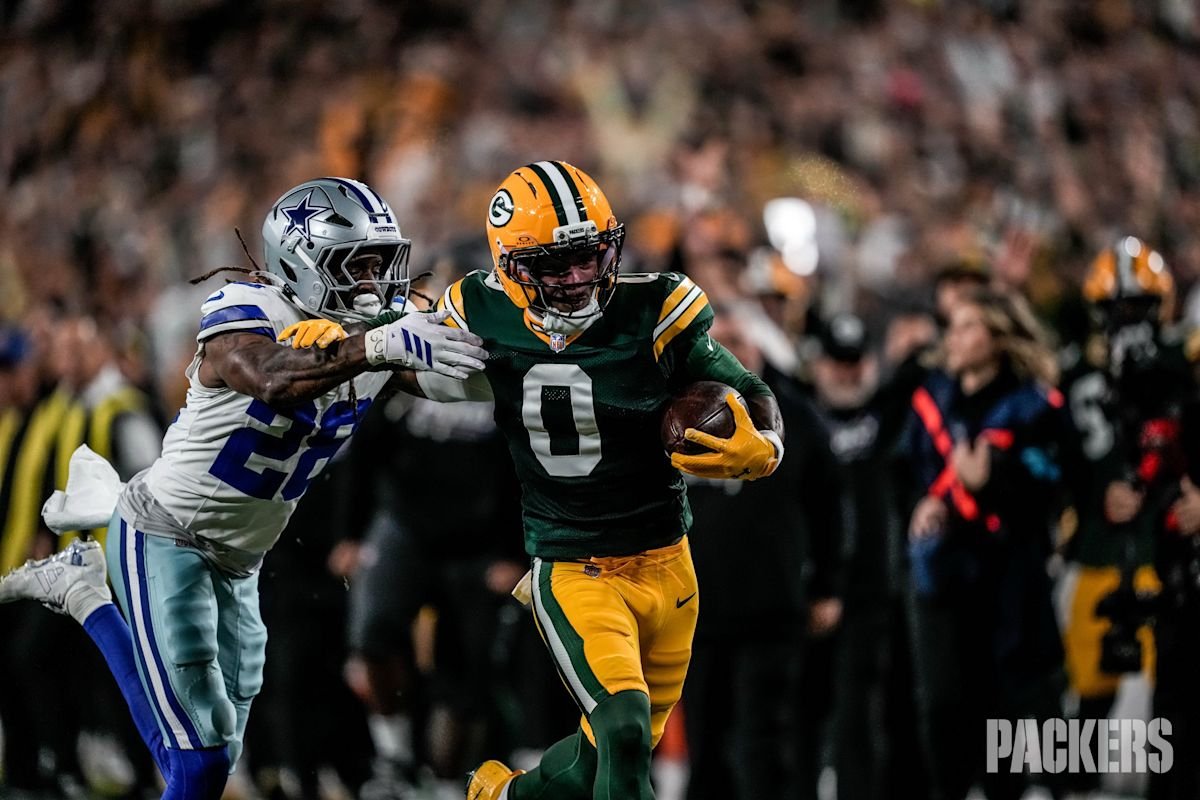 Matthew Golden, de los Green Bay Packers, con la camiseta verde número 0, corre con la pelota bajo el brazo derecho mientras un defensor de los Dallas Cowboys, de blanco con el número 26, intenta frenarlo, con la tribuna de Lambeau Field detrás