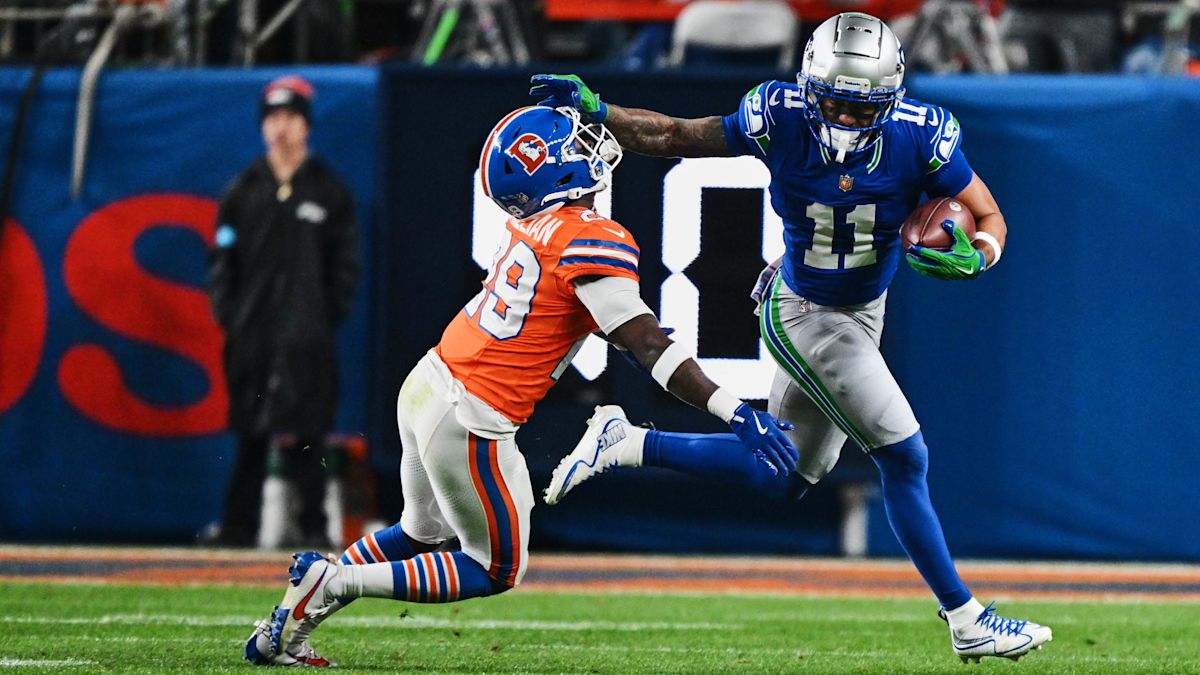 Jaxon Smith-Njigba, con la camiseta azul número 11 de los Seahawks, corre con la pelota y aparta con el brazo a Ja'Quan McMillian, número 29 de los Broncos, en el Empower Field at Mile High