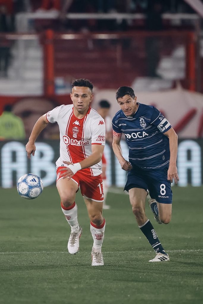 Un jugador de Huracán, con la camiseta blanca y roja, y el capitán de Gimnasia, con la camiseta azul y el número 8, corren detrás de la pelota en el Tomás Adolfo Ducó durante el partido de la fecha 13 (foto: Liga Profesional de Fútbol)