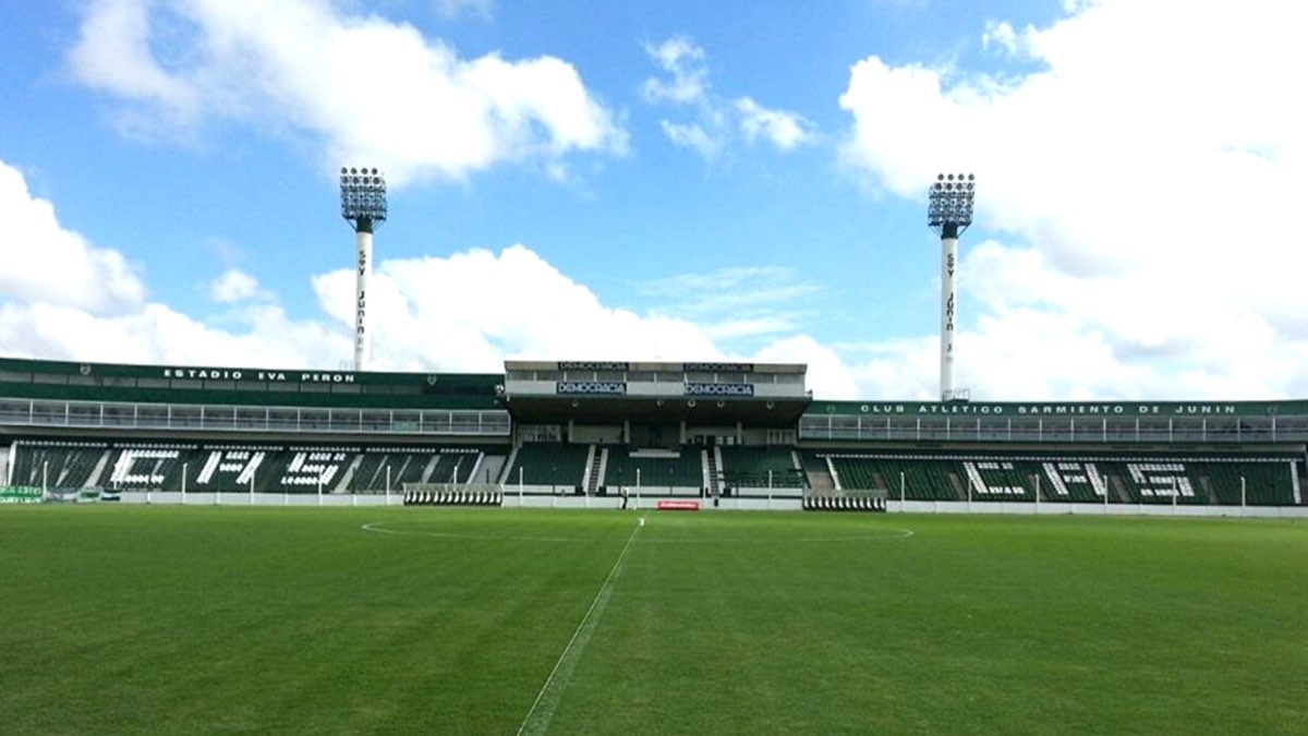 El estadio Eva Perón de Junín, con la platea verde y blanca de Sarmiento, las letras del club en las gradas y dos torres de iluminación bajo un cielo con nubes; foto de archivo de 2015 (foto: Paolazaccardi / Wikimedia Commons, CC0)