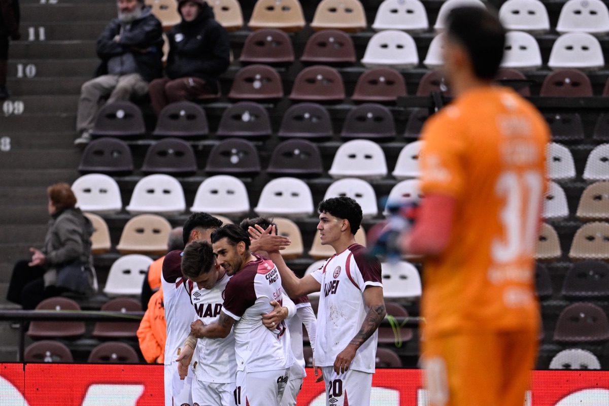 Jugadores de Lanús, de blanco y bordó, se abrazan tras el gol ante Platense, con el arquero local, de naranja, fuera de foco en primer plano (foto: Liga Profesional de Fútbol)