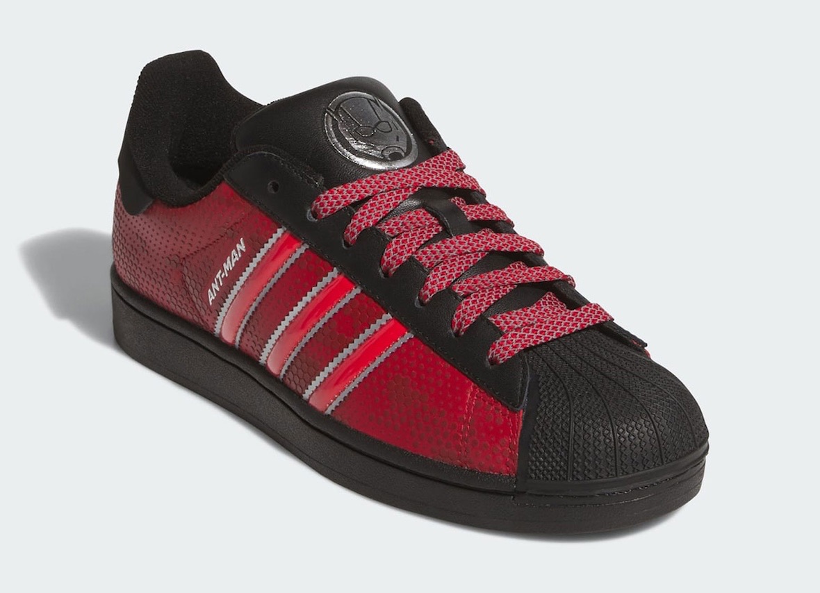 adidas Superstar Ant-Man en vista de tres cuartos con laterales rojos texturados, tiras rojas con bordes plateados, puntera negra y el logo plateado del personaje en la lengüeta