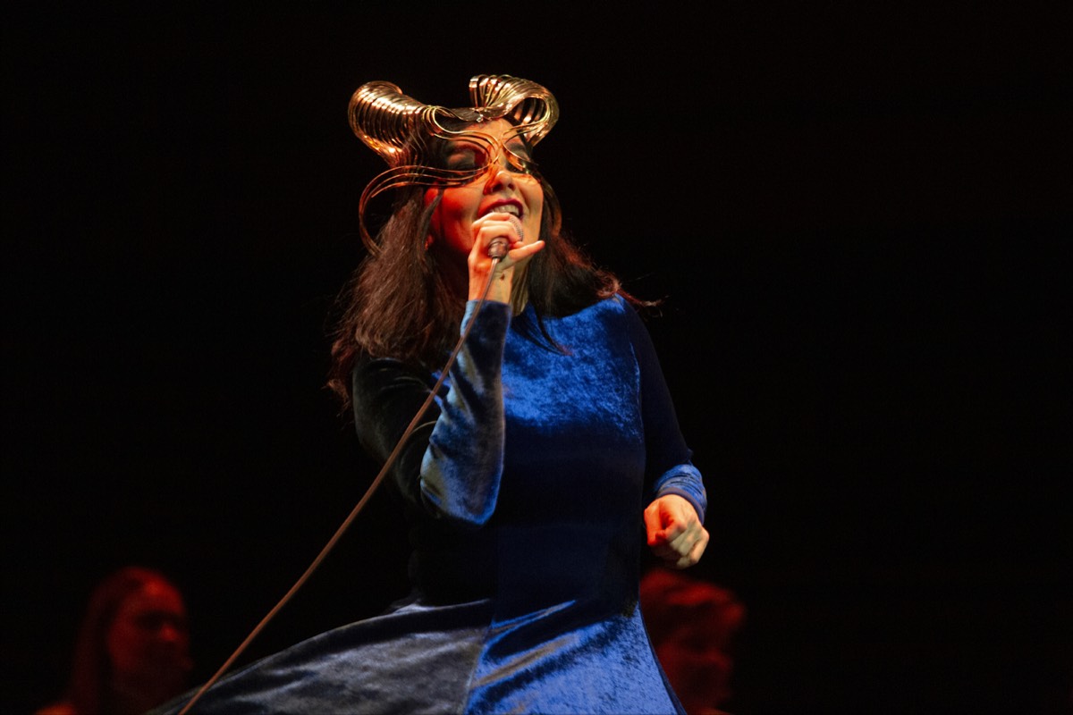 Foto oficial de Björk en el concierto Orkestral del 24 de octubre de 2021: con el vestido de terciopelo azul y el tocado dorado, canta con el micrófono en la mano sobre un fondo oscuro