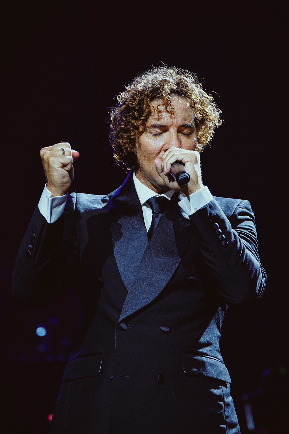 Foto de prensa oficial del show de David Bisbal en el Movistar Arena: el cantante, con el pelo enrulado, traje cruzado oscuro, camisa blanca y corbata, canta con los ojos cerrados tapándose la boca con el micrófono y el otro puño en alto, sobre un fondo negro