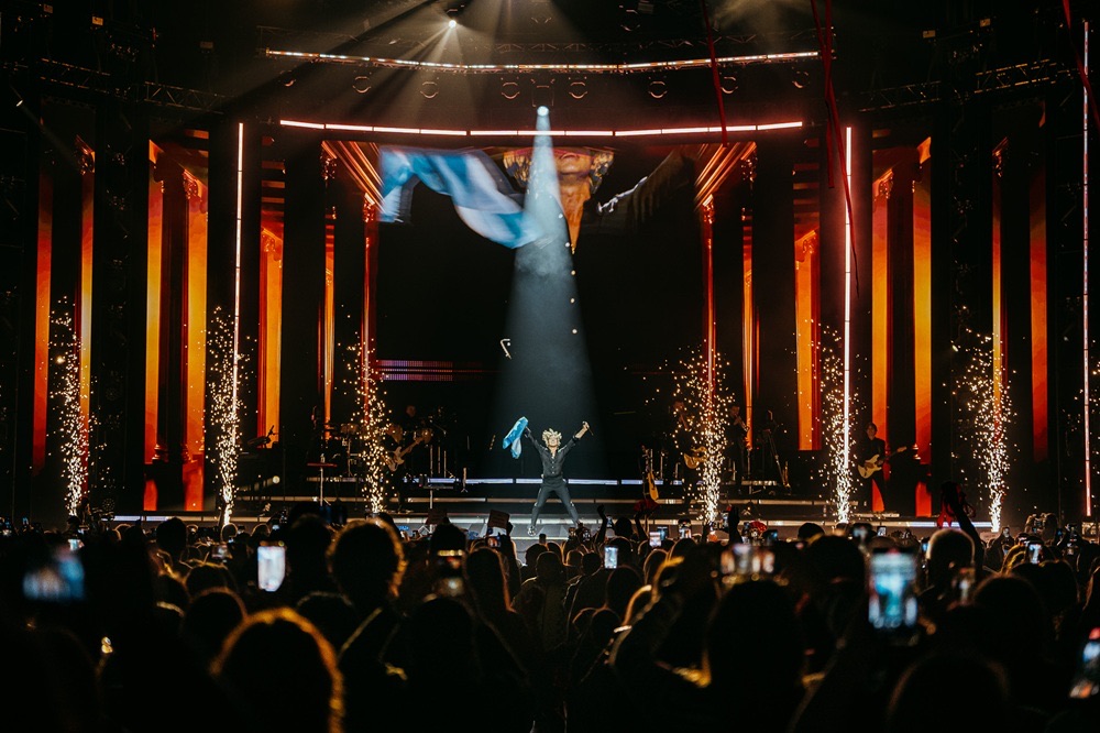 Foto de prensa oficial del show de David Bisbal en el Movistar Arena: el cantante, solo en el centro del escenario bajo un haz de luz, levanta una bandera argentina mientras otra flamea en la pantalla, con columnas iluminadas en naranja, chispas de fuegos fríos a los costados y el público con los celulares en alto en primer plano