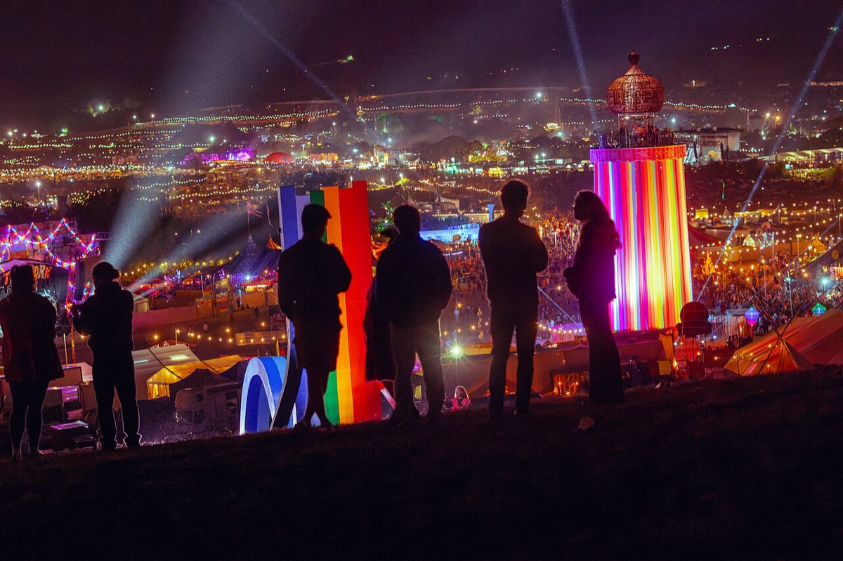 Un grupo de personas a contraluz mira de noche el predio de Glastonbury 2025 desde una colina, con una torre de cintas de colores iluminada, haces de luz y miles de luces de carpas y escenarios de fondo. Foto: Raph_PH, Wikimedia Commons, CC BY 4.0