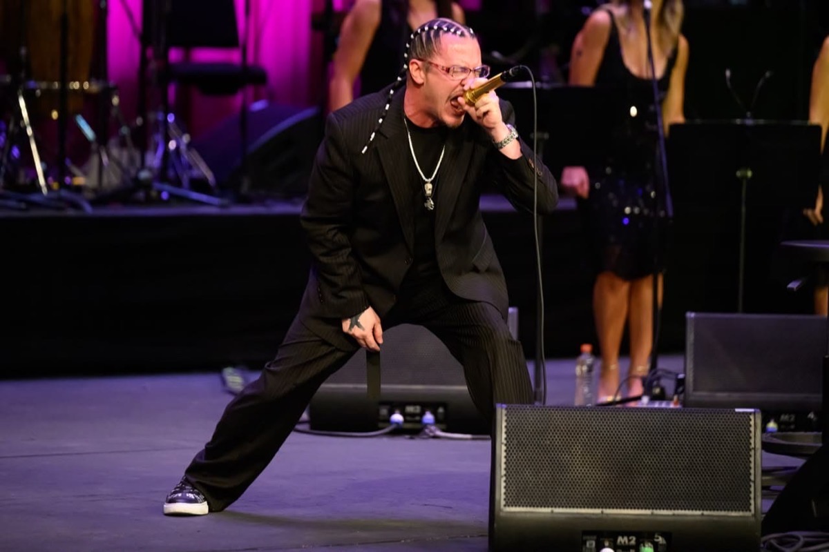Mike Patton con traje negro y trenzas canta con las piernas abiertas y el micrófono dorado pegado a la boca, con las coristas de fondo, en el Palacio de Bellas Artes. Foto: Secretaría de Cultura