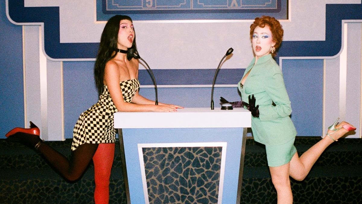 Foto de prensa oficial del remix de "expectations": Olivia Rodrigo, con vestido corto a cuadros blancos y negros, choker y una media roja y otra negra, y Chappell Roan, con traje verde agua, guantes negros y pelo rojo enrulado, posan con una pierna levantada a cada lado de un atril de concurso de TV con dos micrófonos, frente a un decorado azul. Foto: Phoenix Johnson