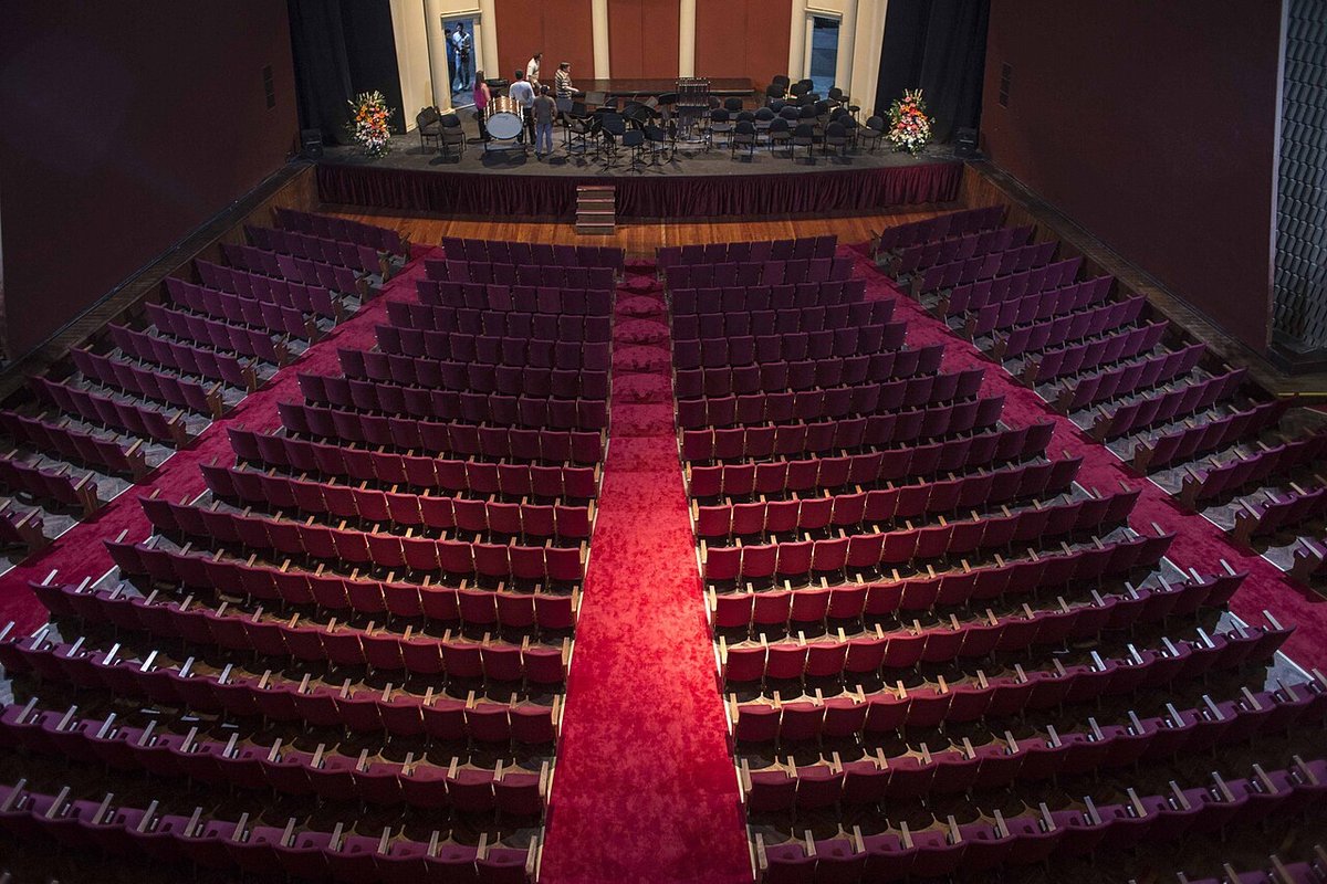 Sala del Teatro Coliseo de Buenos Aires vista desde arriba: filas de butacas bordó a ambos lados de un pasillo central alfombrado en rojo, y al fondo el escenario con atriles, sillas y una batería. Foto: Enrico Fantoni, Wikimedia Commons, CC BY-SA 4.0