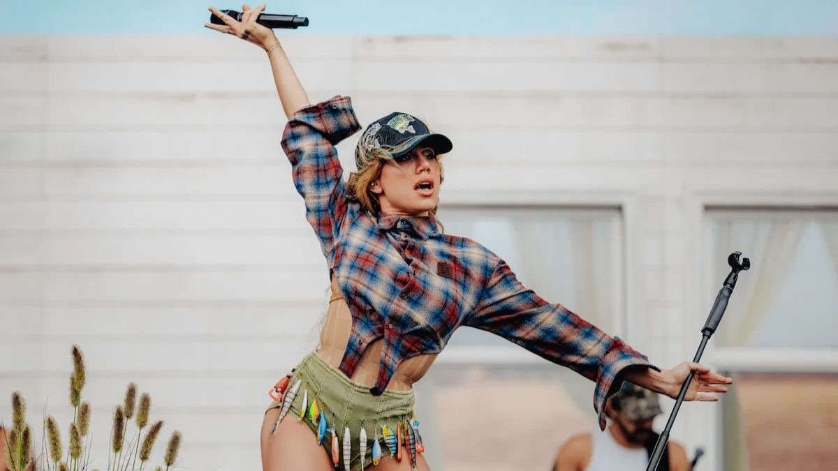 Foto de prensa en vivo de Slayyyter: con gorra, camisa escocesa cortada y un short verde con señuelos de pesca colgando, levanta el micrófono con el brazo estirado frente a una pared de madera blanca. Foto: Joshua Druding
