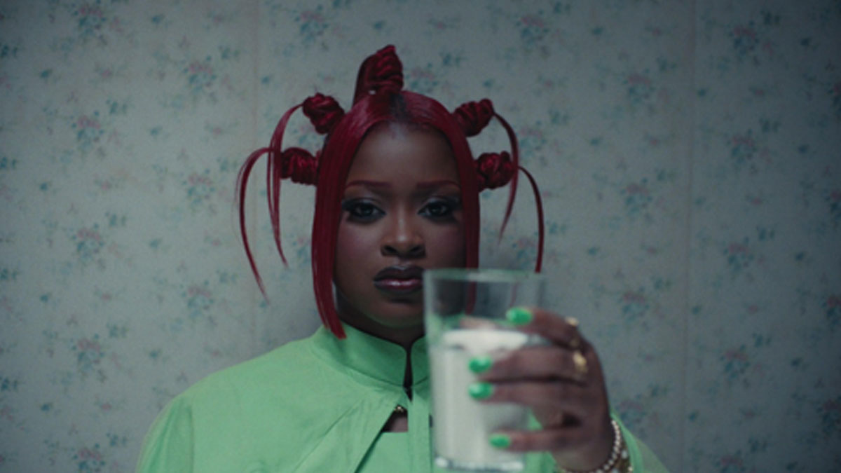 Imagen oficial del video de "CANDLE WAX" (foto de archivo, julio de 2026): Tierra Whack, con el pelo rojo atado en varios rodetes y mechones sueltos, campera verde clara y uñas verdes, mira a cámara y sostiene un vaso con un líquido blanco frente a una pared empapelada con flores