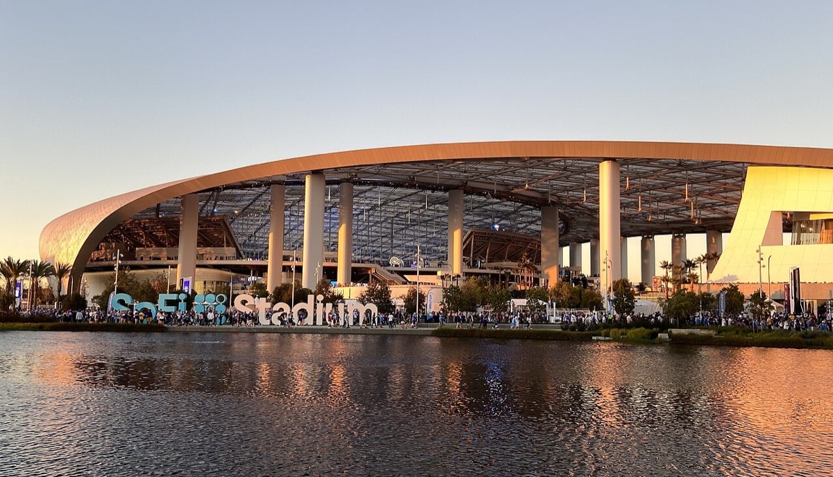 El SoFi Stadium de Inglewood al atardecer, con su techo curvo y translúcido reflejado en un lago y el cartel SoFi Stadium en letras de colores en la entrada. Foto: Troutfarm27 / Wikimedia Commons (CC BY-SA 4.0)