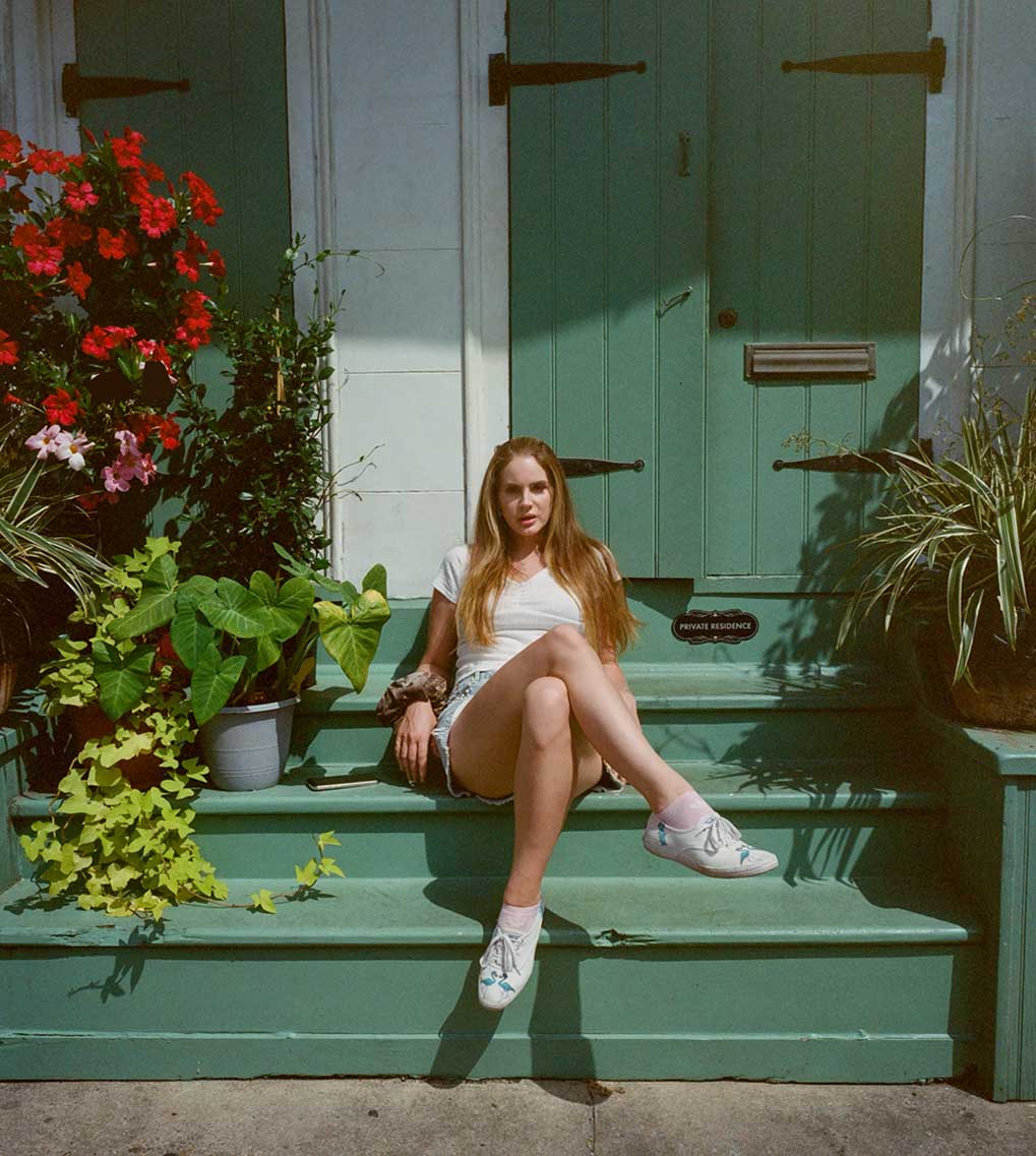 Foto oficial de archivo de Lana Del Rey: sentada en los escalones verdes de la entrada de una casa, con remera blanca, short de jean y zapatillas blancas, entre macetas con flores rojas y plantas. Foto: web oficial de Lana Del Rey