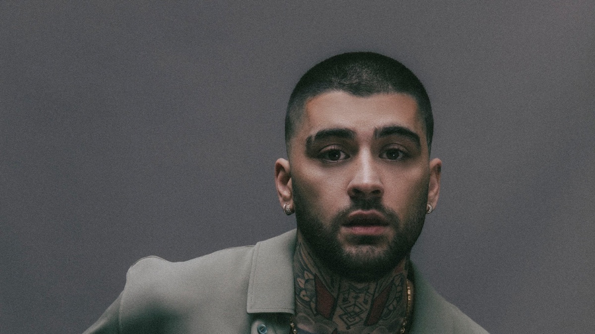 Retrato de prensa de ZAYN con el pelo rapado, barba, aros y tatuajes en el cuello, con camisa verde grisácea frente a una pared gris
