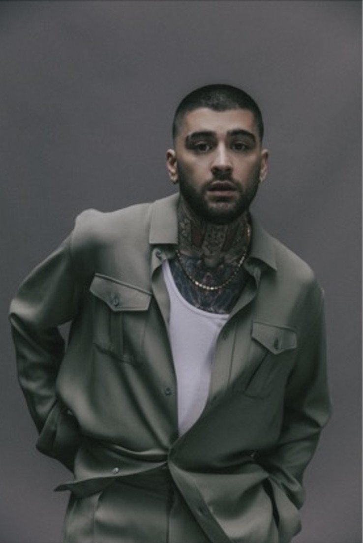 Foto de prensa de ZAYN de cuerpo casi entero, con el pelo rapado, barba y tatuajes en el cuello, camisa verde grisácea abierta sobre una musculosa blanca, cadena dorada y los brazos hacia atrás, frente a un fondo gris