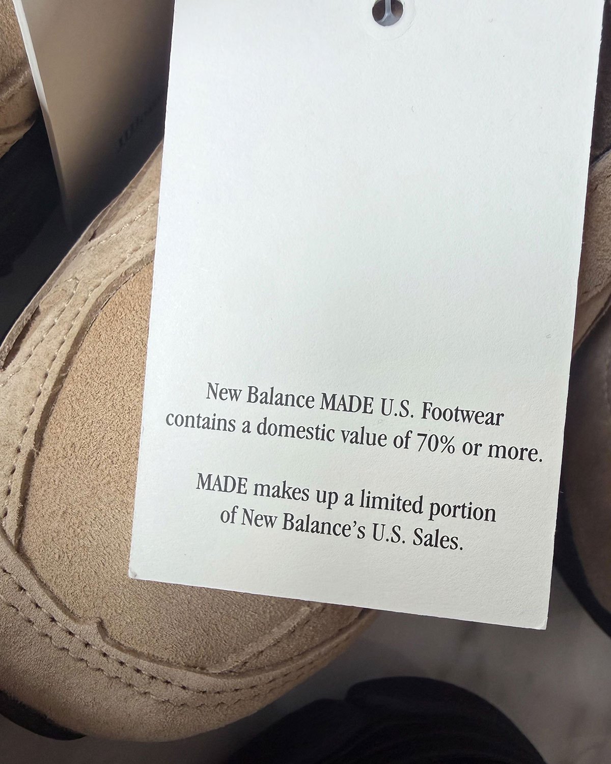 Etiqueta colgante blanca de New Balance MADE U.S. sobre un JJJJound x New Balance 990v3 de gamuza tan, que indica un valor doméstico del 70% o más