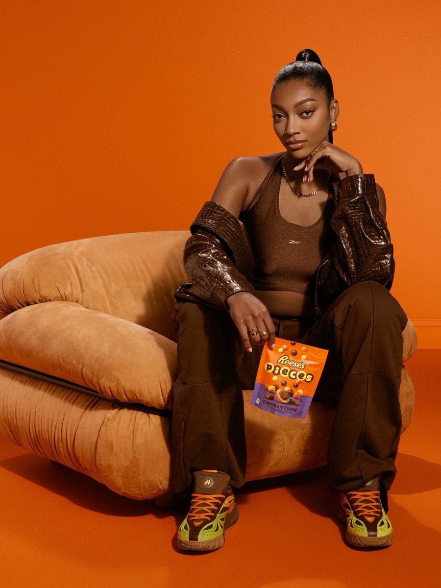Angel Reese sentada en un sillón sobre fondo naranja, vestida de marrón, con un paquete de Reese's Pieces y las Angel Reese 1 Reese's puestas