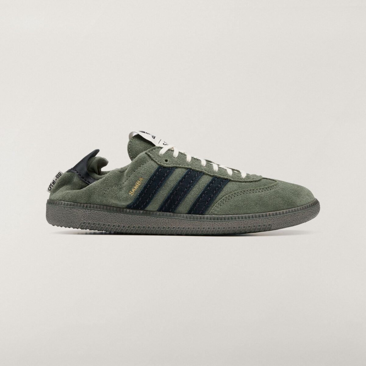 Vista lateral del adidas Samba OG Base Green de Song for the Mute en gamuza verde oliva, con las tres tiras en gamuza azul oscuro, la palabra Samba en dorado, cordones crema, suela verde grisácea y el talón doblado hacia adentro con la etiqueta SFTM-010