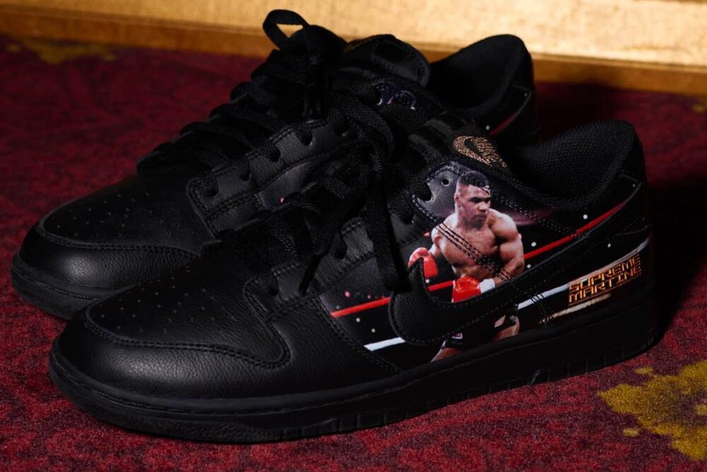 Par del Supreme x Martine Rose x Nike Dunk Low Tyson apoyado sobre una alfombra bordó, en cuero negro con cordones negros, la foto de Mike Tyson con guantes rojos sobre el lateral y las letras doradas Supreme Martine en el talón