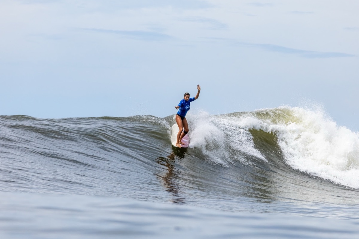 Avalon Gall, con lycra azul, camina sobre su longboard con un brazo en alto en una ola de El Sunzal
