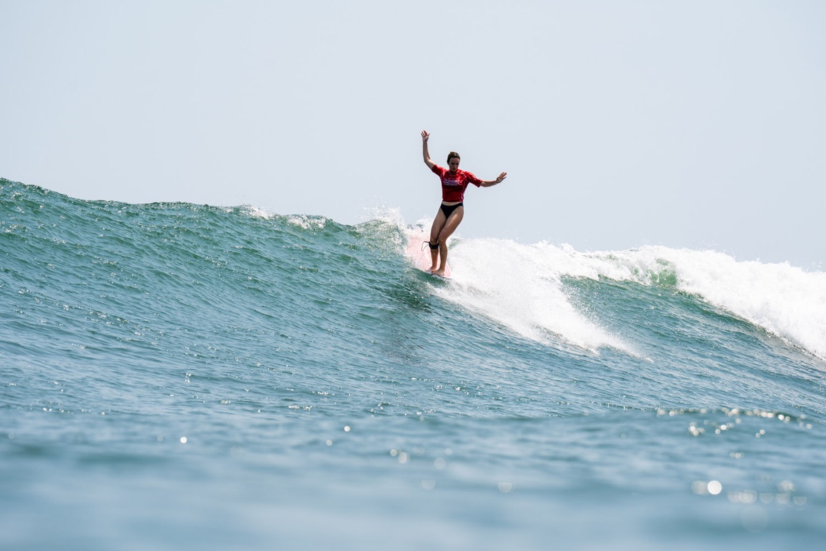 Rachael Tilly, de lycra roja, camina sobre el longboard con los brazos abiertos en el labio de una ola en El Sunzal