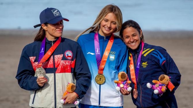 Isabella Assmann, con campera celeste y blanca de la Argentina y la medalla de oro, sonríe en el centro del podio del bodyboard femenino en Playa Grande, junto a una deportista de Chile con la plata y otra de Colombia con el bronce