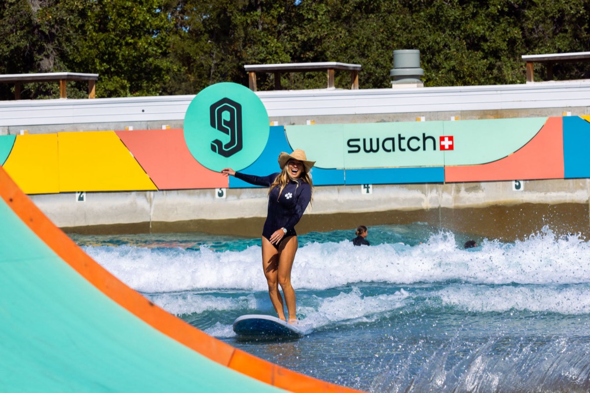 Coco Ho, con traje negro y sombrero de ala ancha, saluda mientras surfea en Waco Surf frente al cartel de Swatch y el logo de los Nines, en la edición 2024