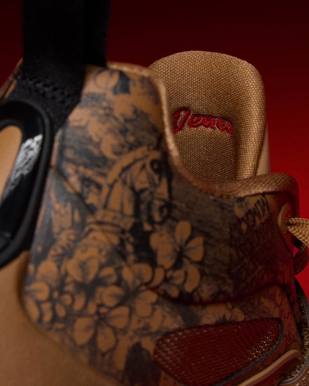 Primer plano del talón del Jordan Tatum 5 Laser con grabados de flores y escenas sobre el material tostado, y el nombre Deuce bordado en rojo detrás de la lengüeta