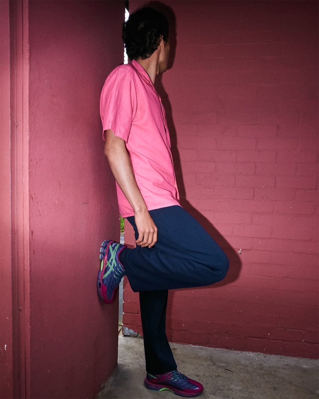 Modelo con camisa rosa y pantalón azul marino apoyado en una pared bordó, con una pierna flexionada contra el muro y la ASICS US8-S GEL-SJ33 Deep Plum/Fresh Grass puesta